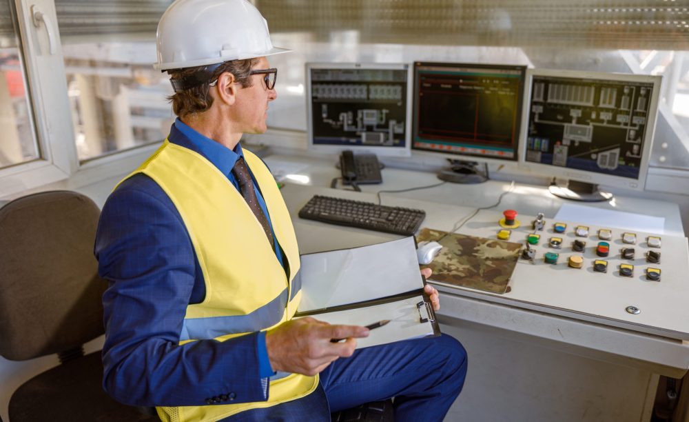 Matured man in work vest and safety helmet sitting at the table with computers and holding papers
